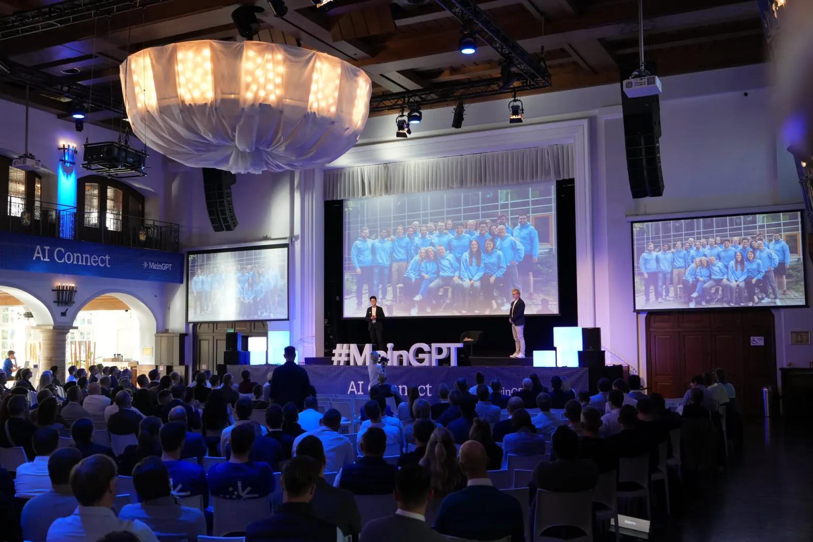 Voller Saal mit Blick auf die Hauptbühne, das Team auf den Leinwänden